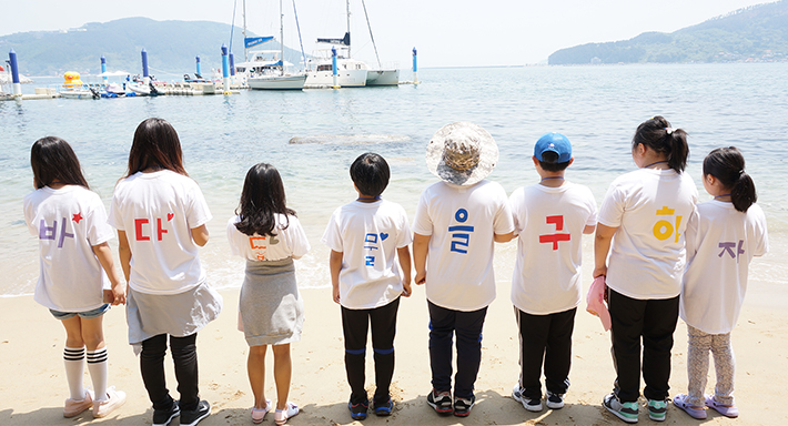 아이들이 등 뒤에 바,다,동,물,을, 구,하,자 가 적힌 티셔츠를 입고 바다 앞에서 찍은 사진 